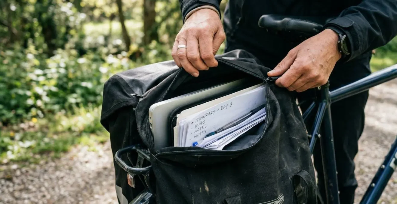 Deux mains ouvrent une sacoche porte-bagages noire révélant un ordinateur portable et des documents rangés à l'intérieur